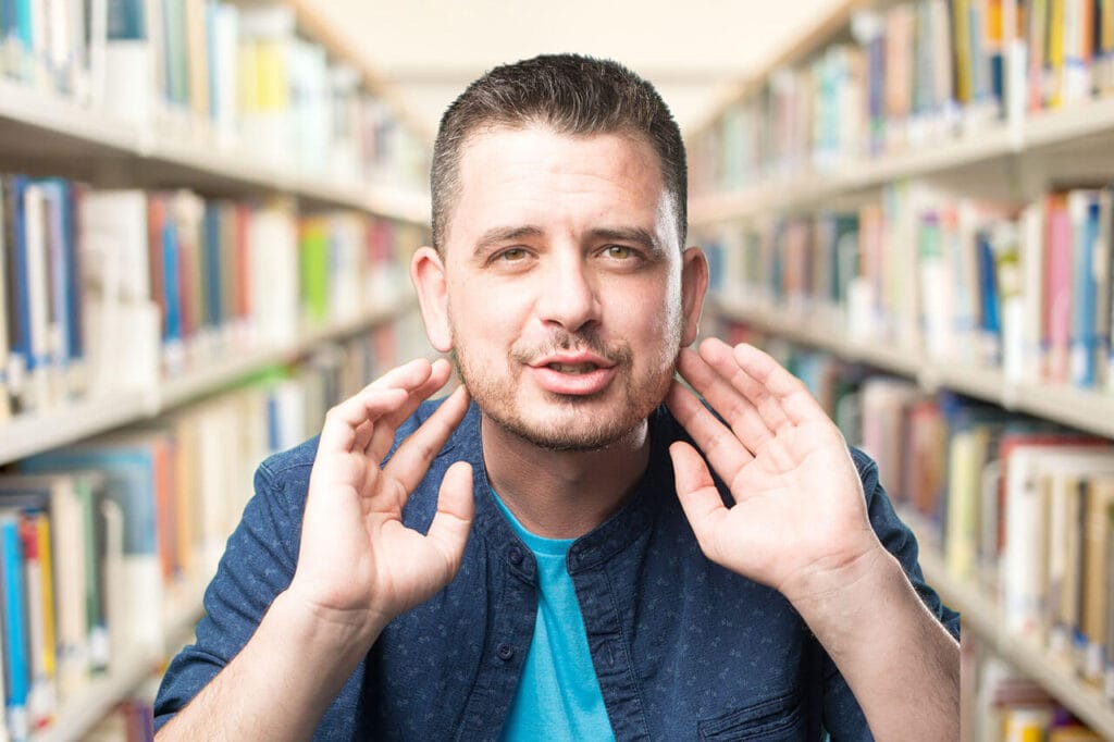 Homem concentrado em uma biblioteca, fazendo gesto de escutar atentamente, simbolizando o aprendizado de expressões idiomáticas em inglês.